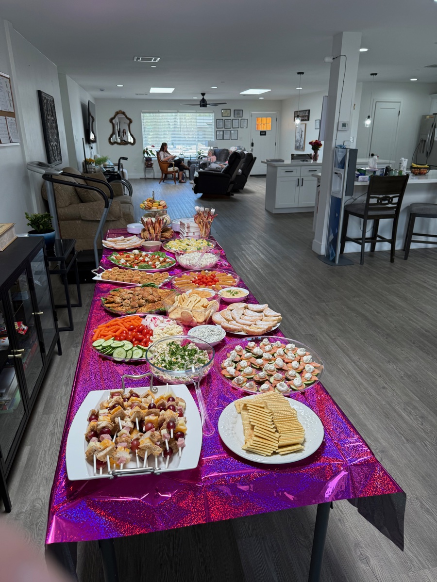 Long buffet table set with appetizers, finger sandwiches, and desserts on a pink tablecloth in the open dining area