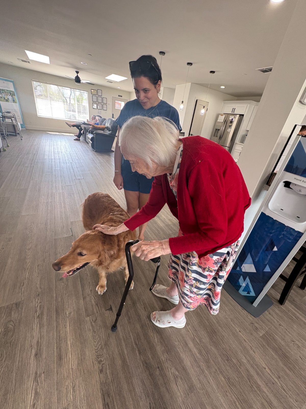 A resident in a red cardigan leans on her cane to pet Maui, with his handler smiling beside them